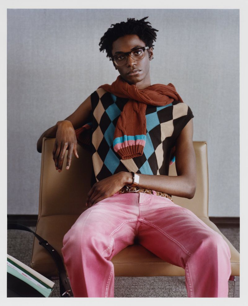 Portrait of a young Black man in pink pants and a patterned sweater vest, seated on a chair in a minimalist studio.