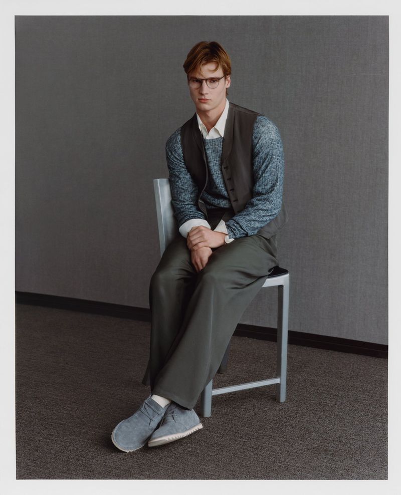 A fair-skinned, brown-haired adult male model wearing glasses, a vest, long-sleeved sweater, and pants sitting on a metal chair in a studio.