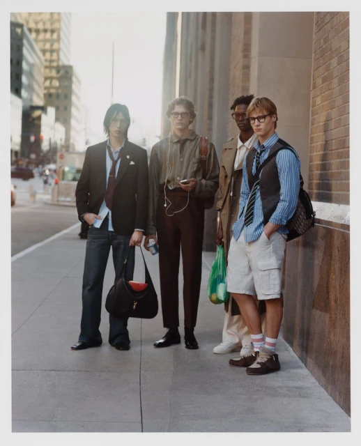 Four fashionable young men stand on a city sidewalk in casual-yet-stylish attire, posing against a blurred backdrop of city buildings.