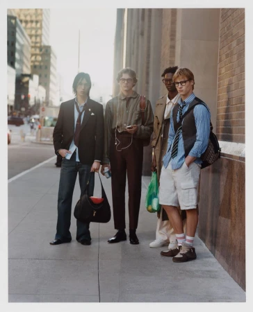 Four fashionable young men stand on a city sidewalk in casual-yet-stylish attire, posing against a blurred backdrop of city buildings.