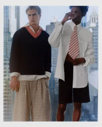 Two male models in tailored menswear standing by a high-rise window overlooking city skyscrapers.