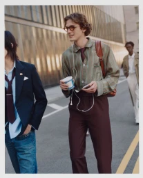 Man wearing a striped shirt and brown trousers walking on a street holding a coffee cup, photographed for Vogue China Men.