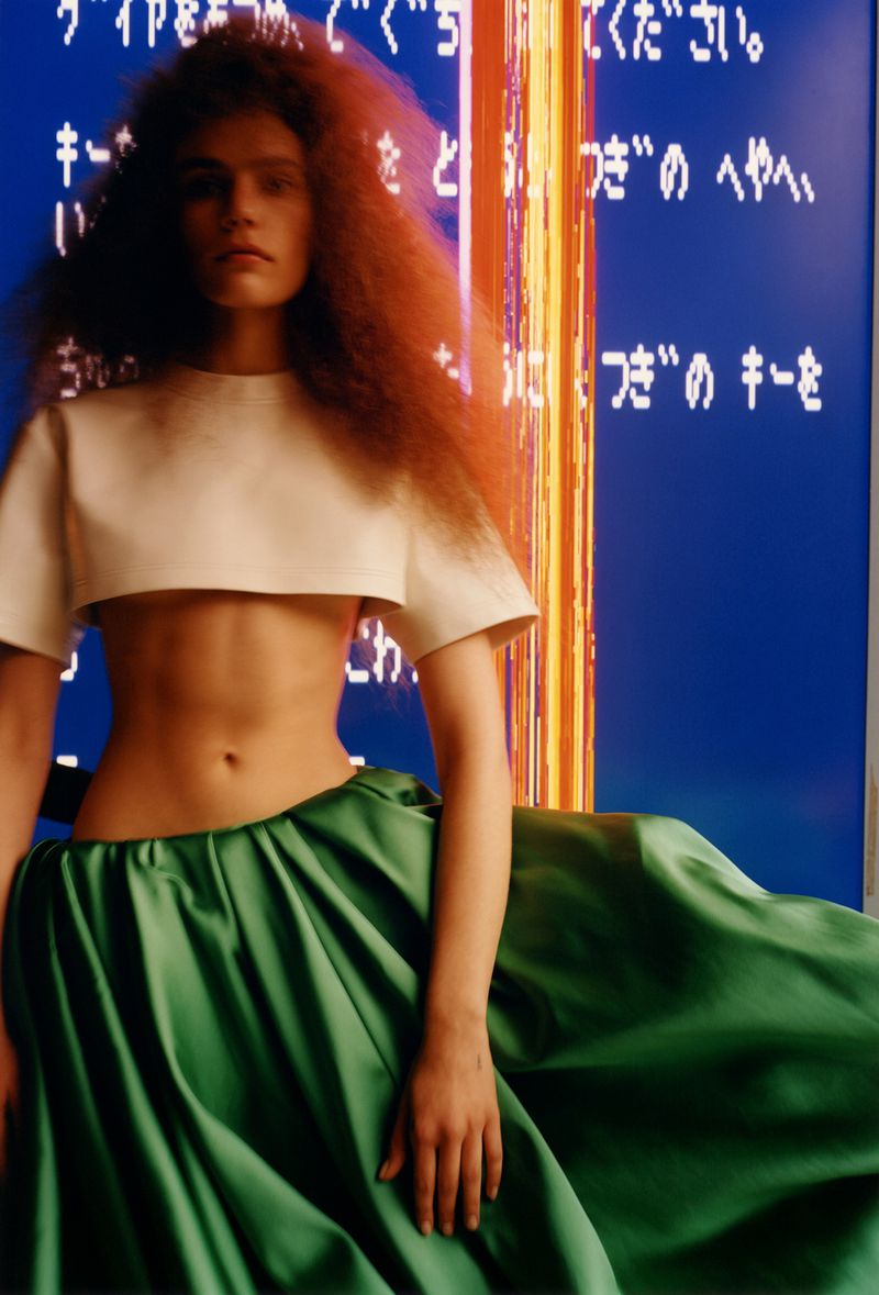 A woman with red hair wears a white crop top and green skirt against a blue background with vertical lights and Japanese text.