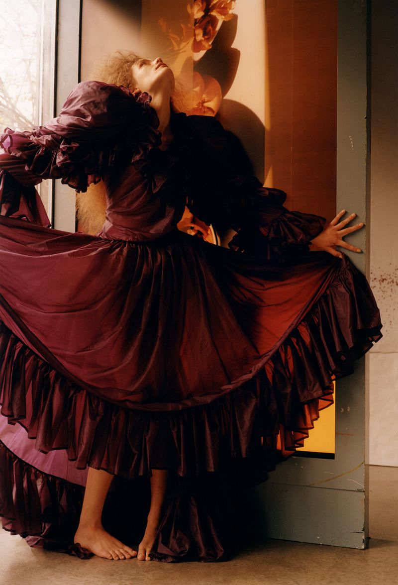 Woman in a voluminous magenta gown poses with outstretched arms near a textured peach wall, exhibiting fashion and photographic artistry.