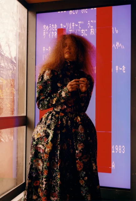 Woman in elaborate floral dress and voluminous auburn hair poses by window with translucent monitor displaying Japanese text and red stripes.