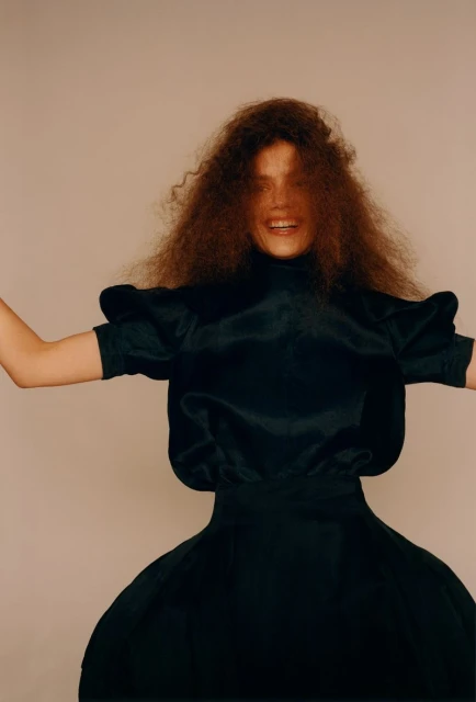 Woman with large curly hair in dark voluminous dress with puffy sleeves smiles with arms spread in a studio shot.