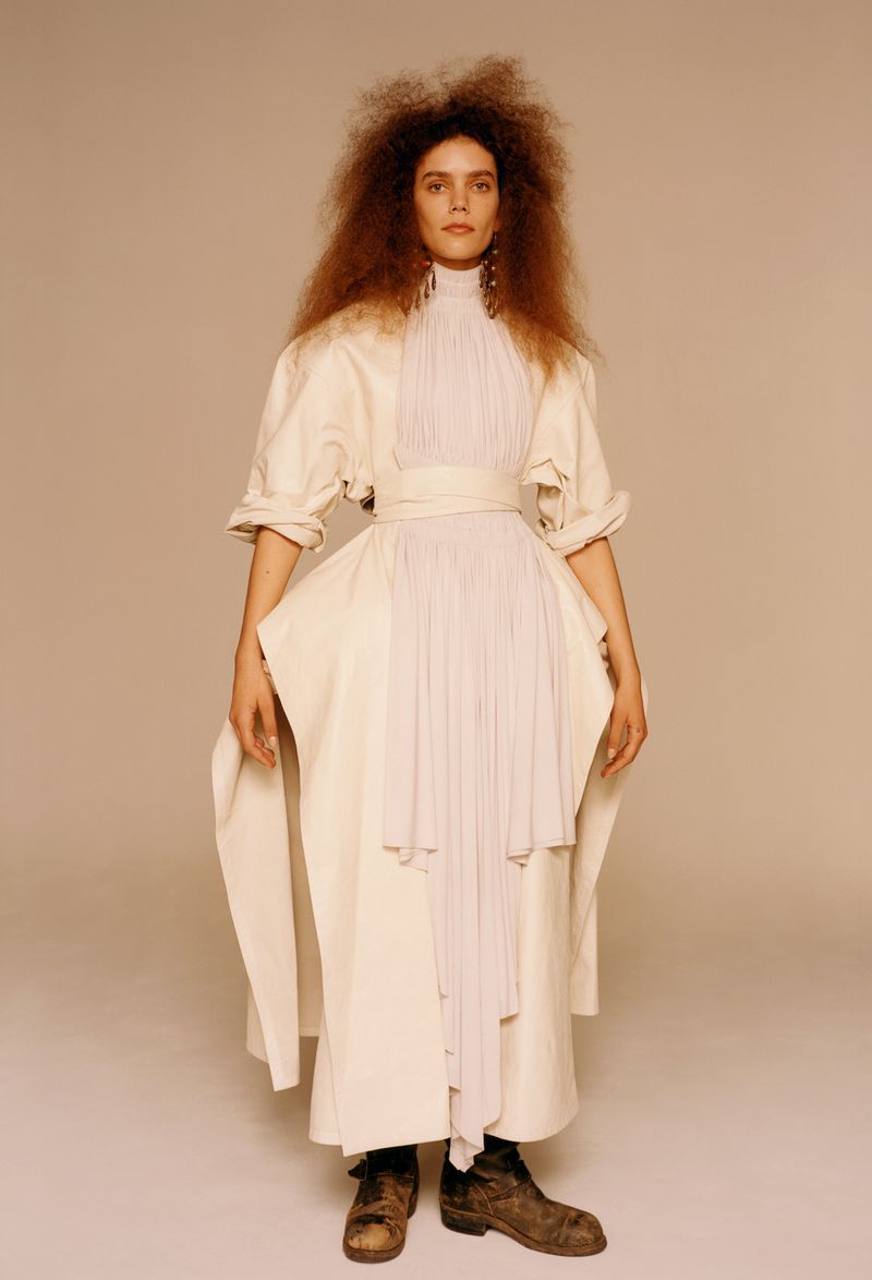 Full-length shot of a model in an off-white dress with a pleated bodice and dark boots posing against a neutral background in studio lighting.
