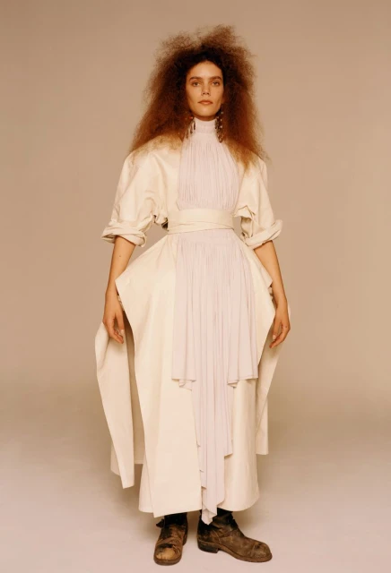 Full-length shot of a model in an off-white dress with a pleated bodice and dark boots posing against a neutral background in studio lighting.