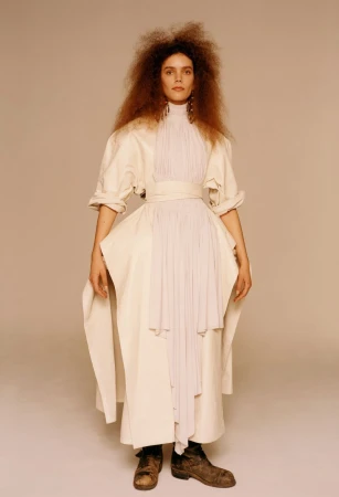 Full-length shot of a model in an off-white dress with a pleated bodice and dark boots posing against a neutral background in studio lighting.