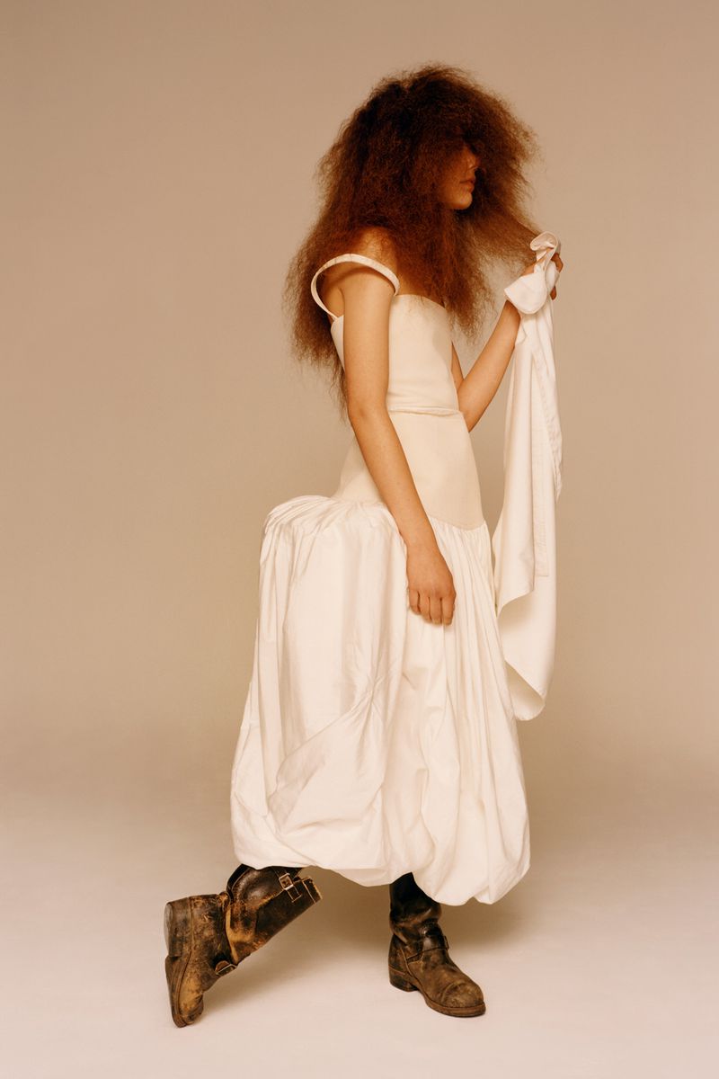 A person poses in a white dress and worn work boots in a studio shot, featuring textured hair and neutral color grading for a fashion editorial.