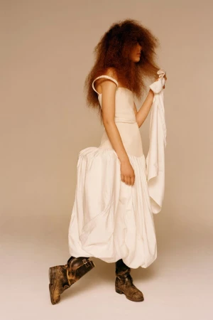 A person poses in a white dress and worn work boots in a studio shot, featuring textured hair and neutral color grading for a fashion editorial.