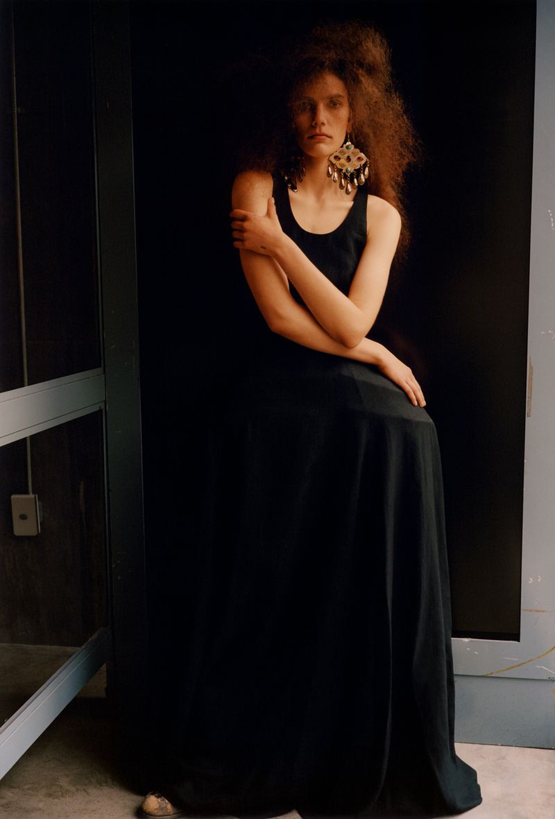 Woman in a black dress poses in front of a dark background. She has large dangling earrings and long curly red hair.