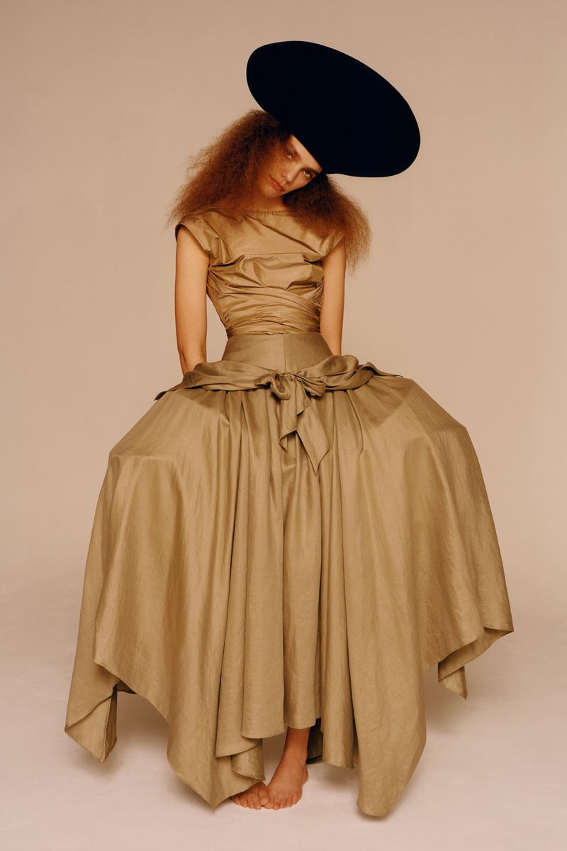 Woman in a light brown dress and black hat seated against a light background, creating a striking fashion editorial image.