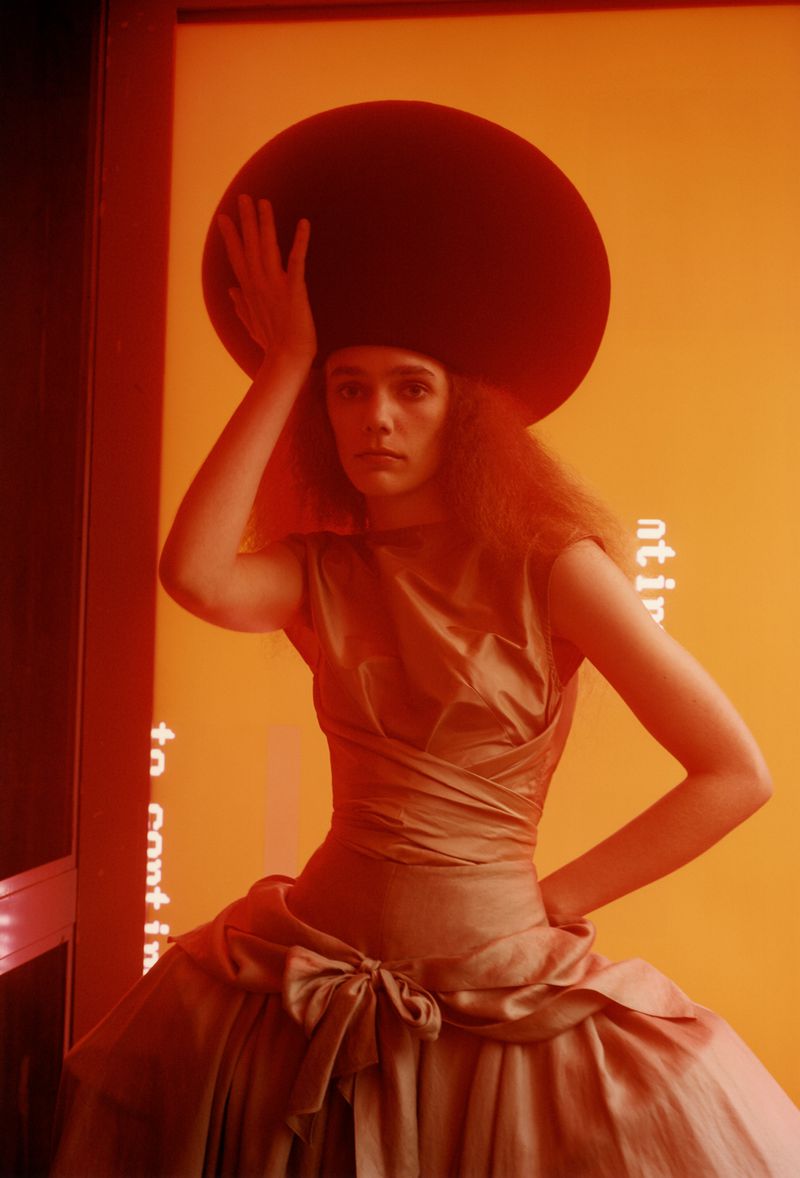 A woman in a golden dress and large hat poses in front of an orange wall with text, illuminated by studio lighting, creating a dramatic fashion shot.