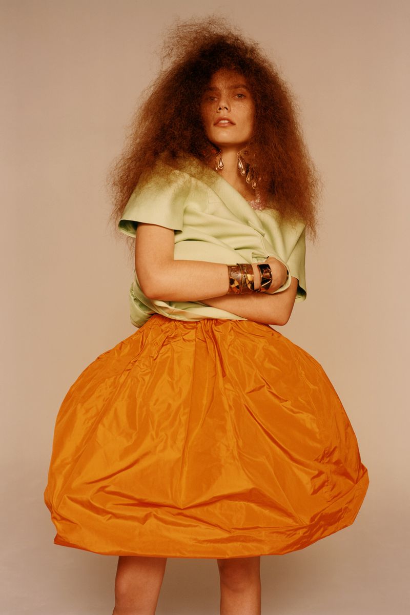 A woman with voluminous afro-style hair poses in a light green top and large orange skirt in a studio environment against a neutral backdrop.