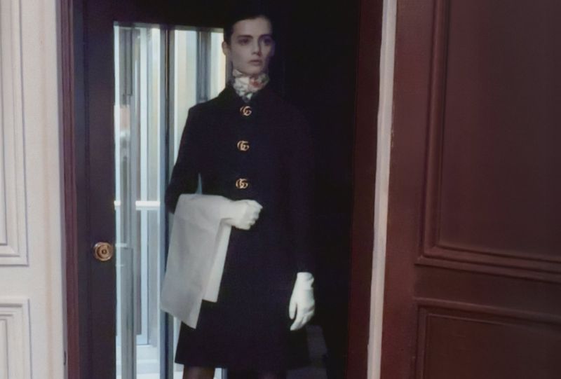 Adult female model in a dark overcoat with white gloves and scarf, posing in a doorway with white door trim and a dark door.
