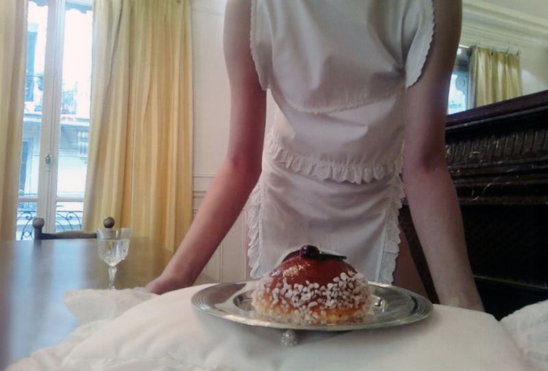 Woman in a white uniform apron stands indoors with a dessert on a silver platter, a glass, and a piano in the background.