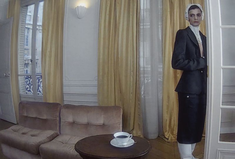 Indoor scene with an adult female fashion model in a dark business suit, standing near a doorway with a cup of coffee on a table.