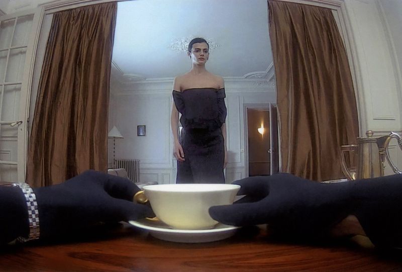 A woman in a black dress stands in an ornate room as a person in gloves holds a steaming teacup, creating a surreal and stylish scene.