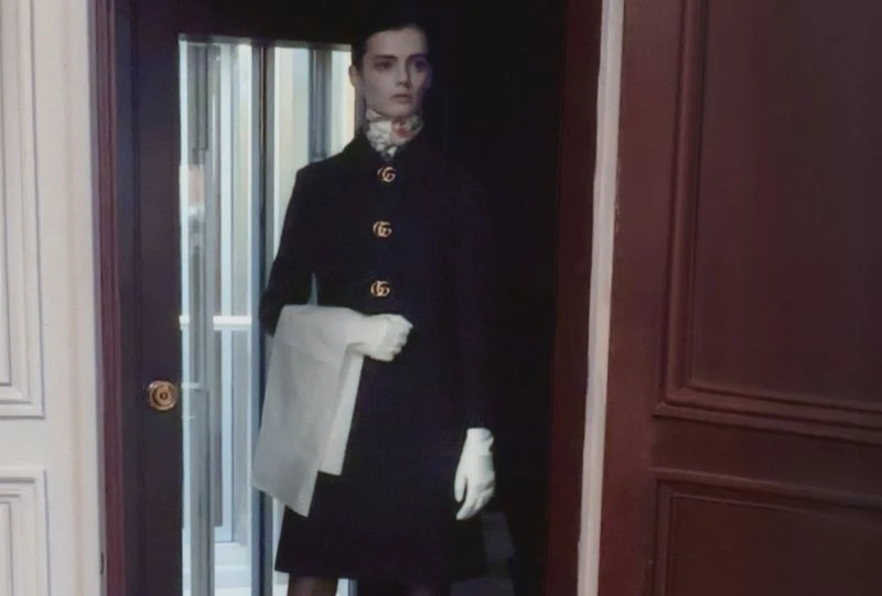 Model Sihana Shalaj wearing a dark coat and white gloves standing inside an interior hallway for AnOther Magazine.