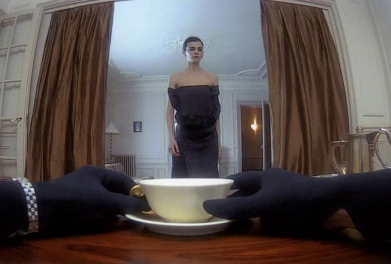 First-person view of black-gloved hands beside a teacup on a table, with model Sihana Shalaj standing in a black dress in the room beyond.