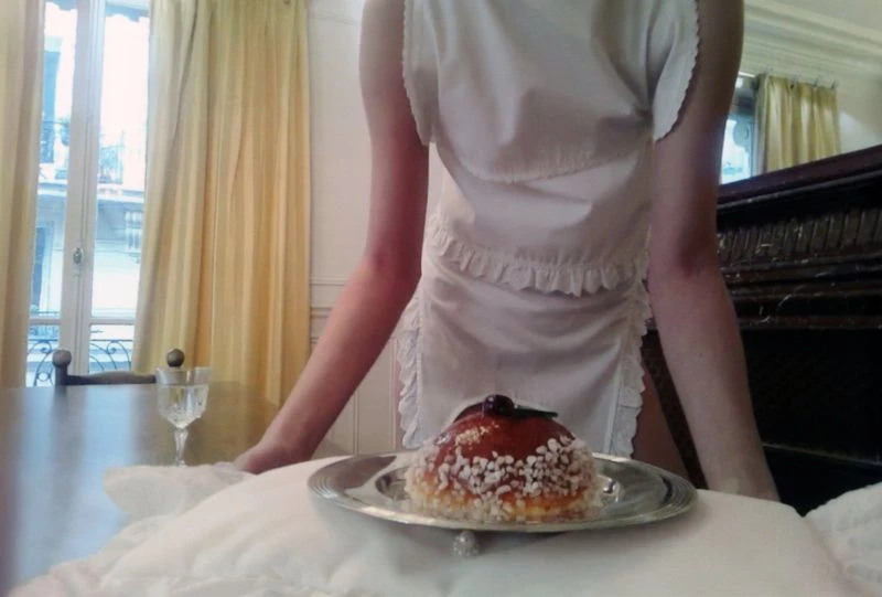 A person wearing a white ruffled apron dress leans forward over a silver platter holding a glazed pastry garnished with a cherry.