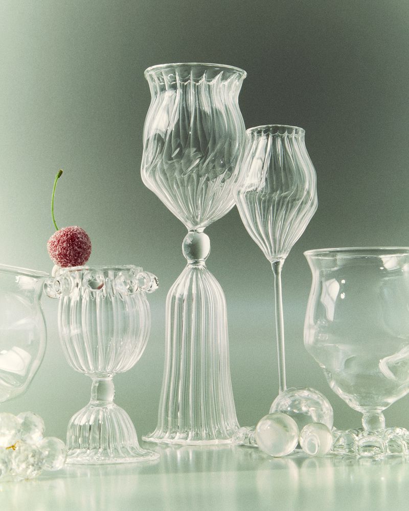 Elegant glassware composition with a variety of clear, ornate glass goblets and a frosted cherry on a reflective surface, set against a soft grey background.