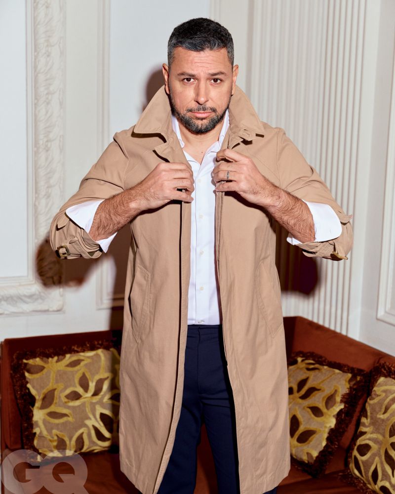 Portrait of an adult male wearing a beige trench coat over a white shirt and dark pants, posing indoors with neutral wall background.