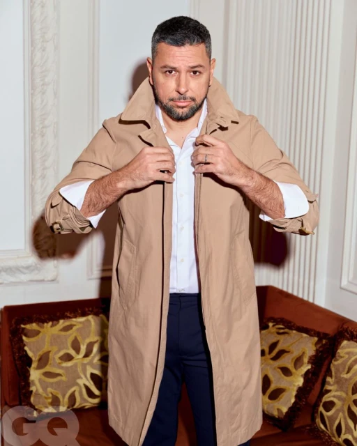 Portrait of an adult male wearing a beige trench coat over a white shirt and dark pants, posing indoors with neutral wall background.