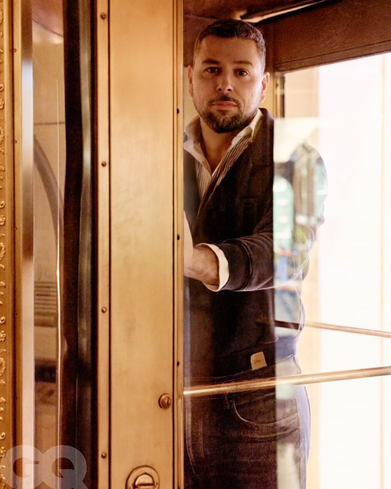 Portrait of an adult man in a jacket and jeans inside an elevator with gold trim and glass doors, looking directly at the camera.