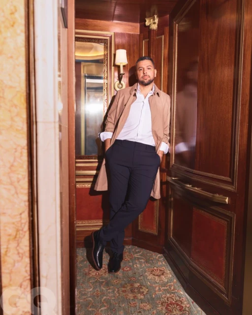 Portrait of adult male posing in beige trench coat, white shirt, and black trousers in hallway with patterned carpet and wood panelling.