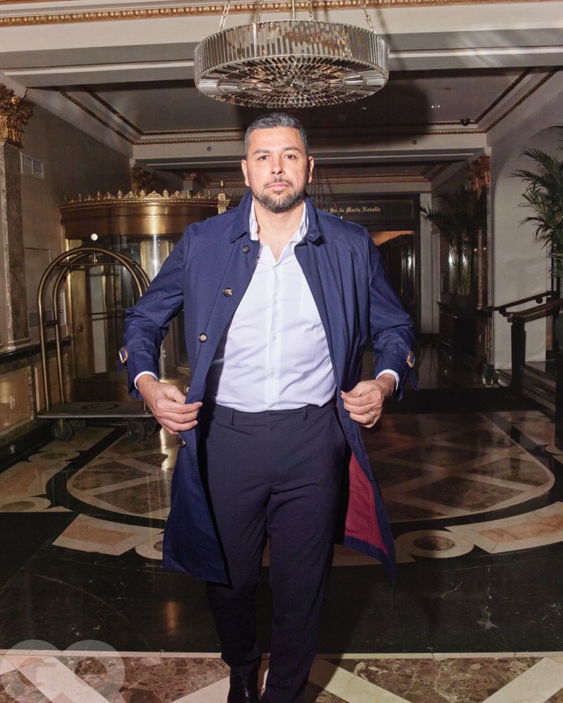 Man in a navy trench coat stands indoors in front of revolving doors. Marble patterned floor and gold architectural details visible.