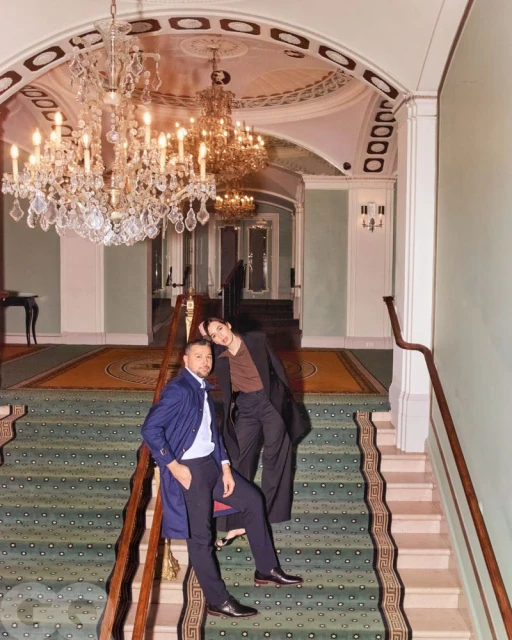 Stylish couple poses on stairs in a grand hall beneath crystal chandeliers. The man wears a blue jacket, and the woman wears a long coat.