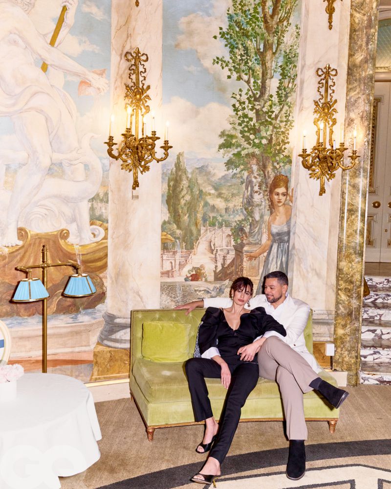 A couple poses in an ornate interior setting with a mural; the woman wears black while the man wears white with tan pants.
