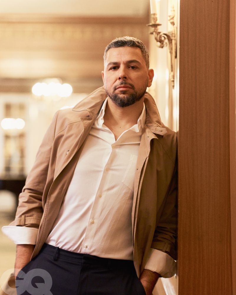 Portrait of a man indoors with a trench coat, white shirt, and dark pants, leaning against a wall with decorative molding and sconces.