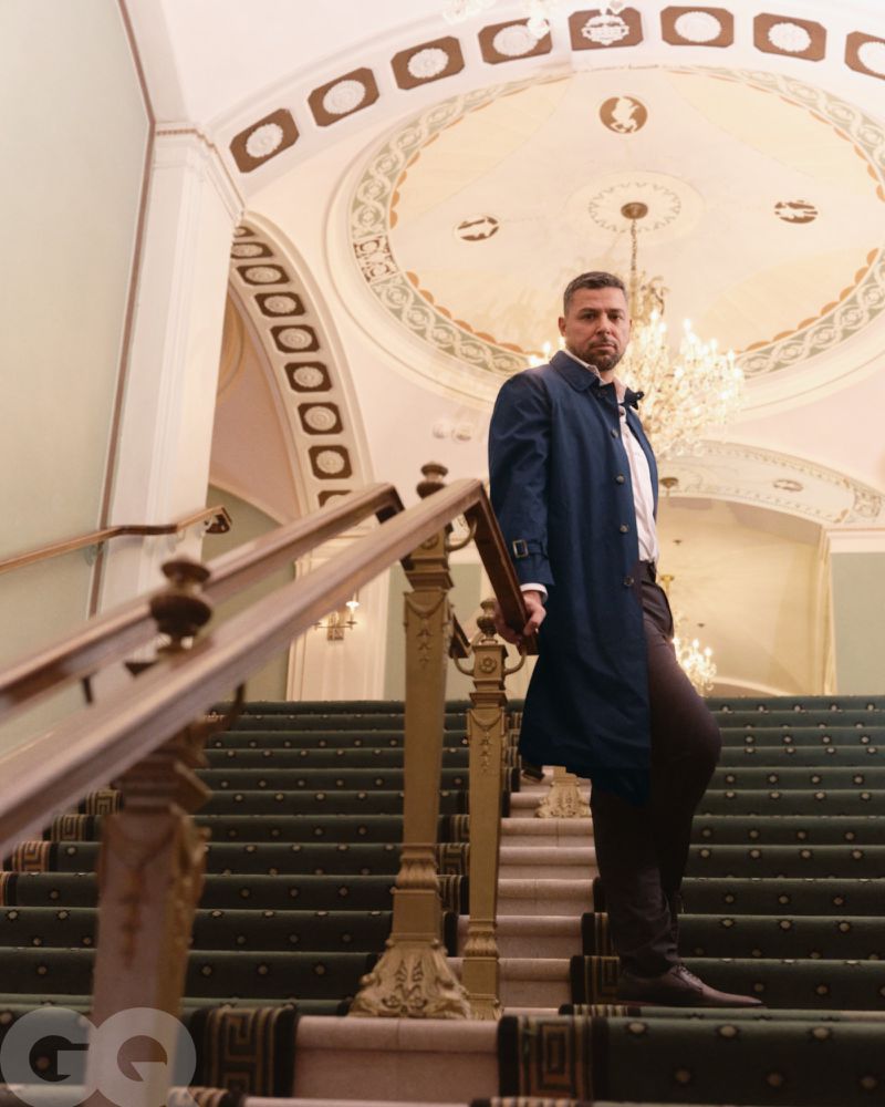 Man in a dark blue trench coat stands on an ornate staircase in an elegant building, gazing directly at the camera with a neutral expression.