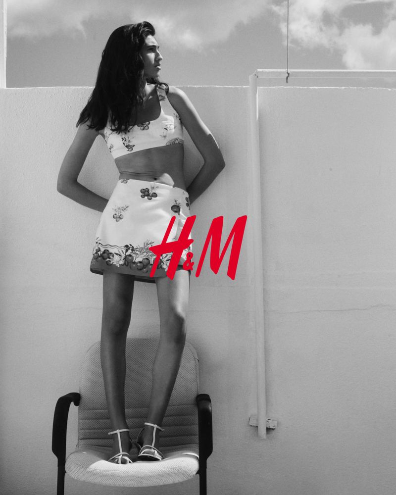 Fashion photo of a female model in a floral crop top and skirt standing on a chair in a black and white outdoor setting.