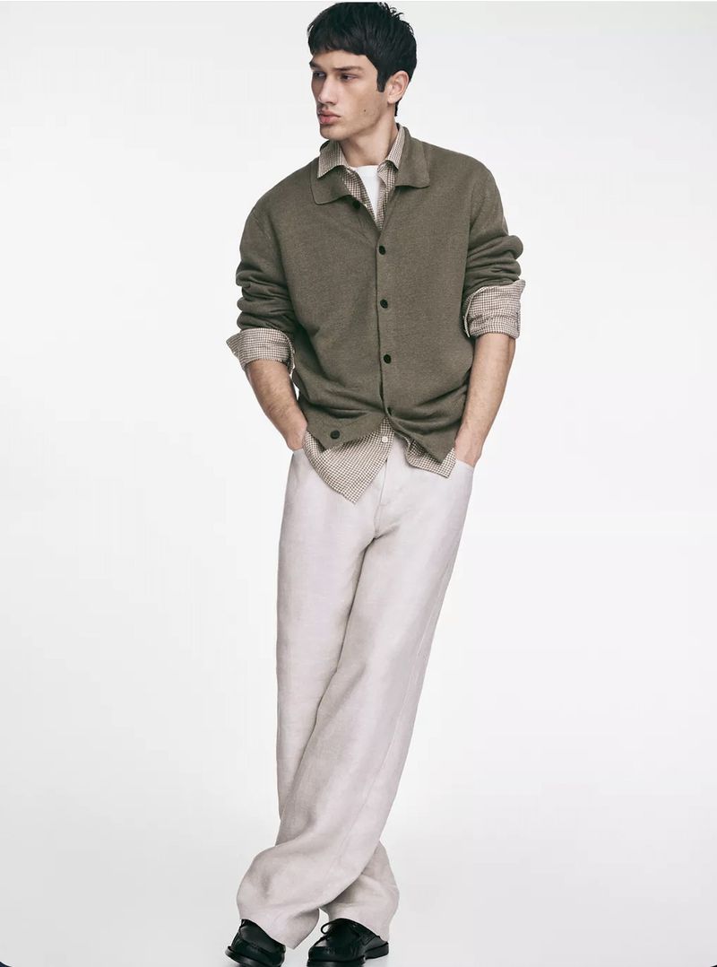Male fashion model posing in a collared button-down shirt and light beige trousers, hands in pockets, against a white backdrop.
