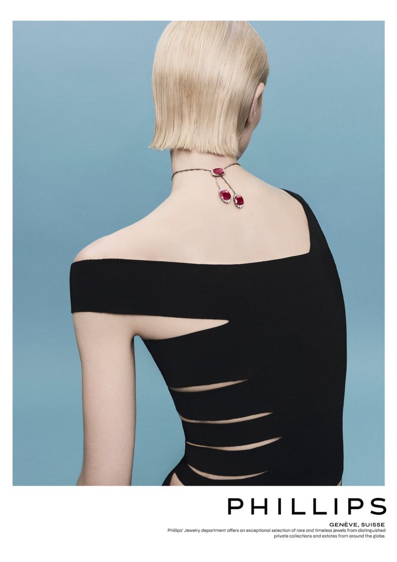 Back view of a female model with short blonde hair wearing a black cut-out dress and a red cherry pendant necklace on a blue background.