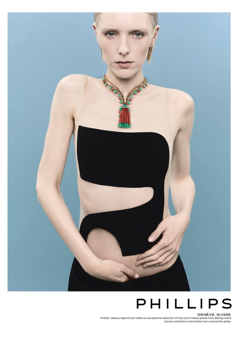 A fair-skinned fashion model poses in a black cutout dress against a light blue backdrop, showcasing an elaborate multi-colored statement necklace.