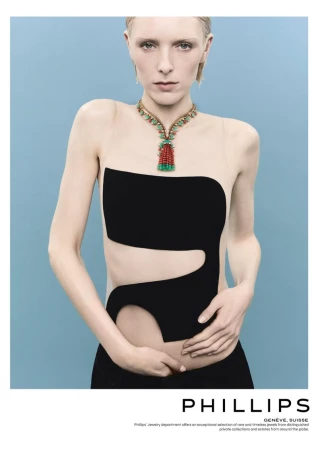 A fair-skinned fashion model poses in a black cutout dress against a light blue backdrop, showcasing an elaborate multi-colored statement necklace.