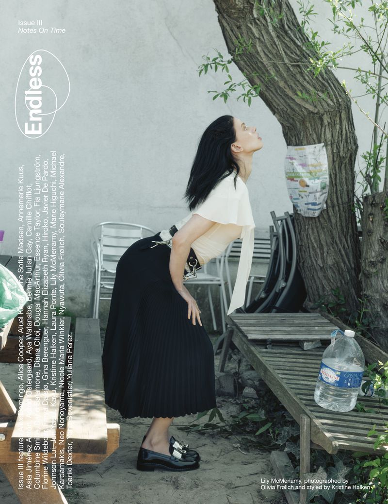 Adult woman in a fashionable midi skirt and top, poses next to a tree and a white wall in an outdoor editorial fashion shot.