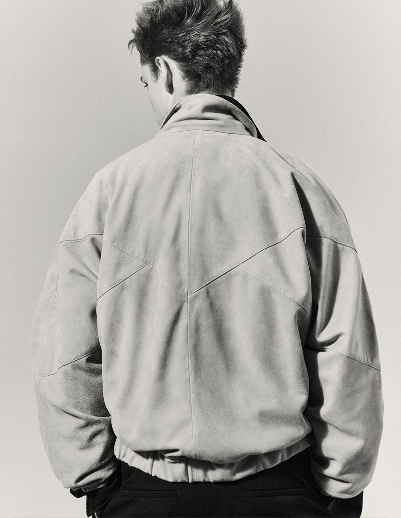 Back view of a person wearing a beige suede jacket with black pants and dark undershirt standing in a neutral-toned studio.