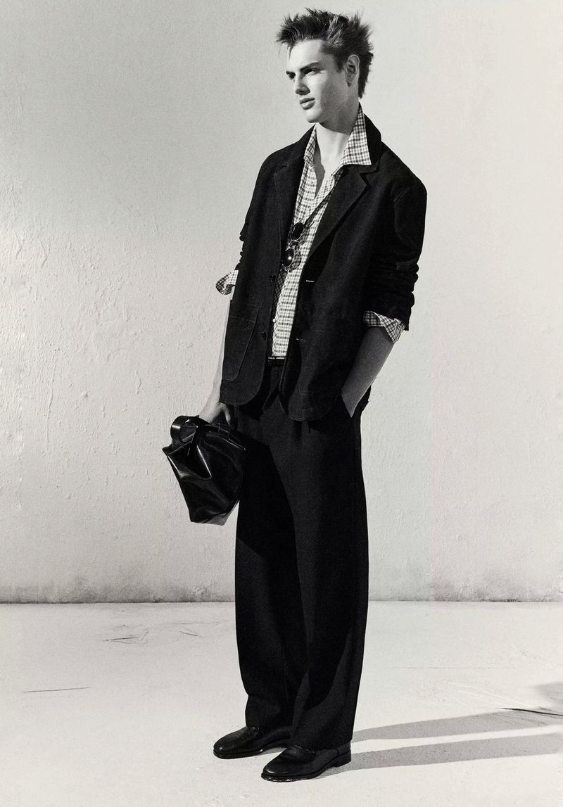 Black-and-white portrait of a young adult male model in a blazer, dress shirt, and slacks, carrying a black leather bag in a photo studio.