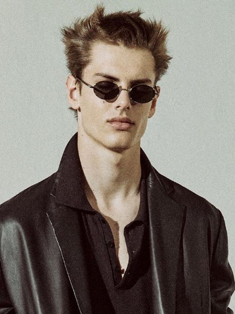 Stylish portrait of an adult male model wearing small oval sunglasses and a black outfit against a neutral backdrop in a photo studio.