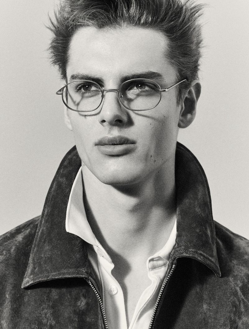 Close-up portrait of a young adult male wearing glasses and a brown suede jacket against a neutral background in black and white.
