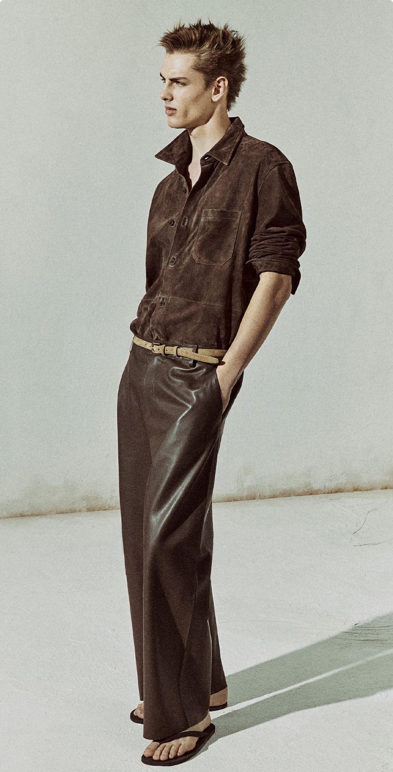 Male fashion model poses in a brown shirt and leather pants against a light background, showcasing a modern, relaxed style.
