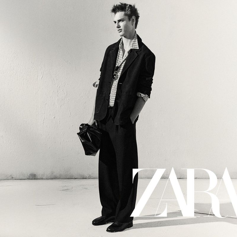 A male fashion model poses in a black suit with a patterned shirt, holding a black bag in a studio with a white backdrop.
