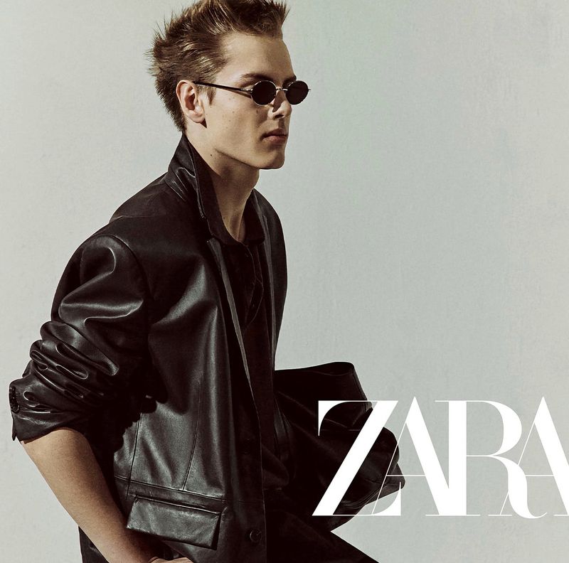 Fashion portrait of a blonde male model in a black leather jacket and small sunglasses, against a light grey background.