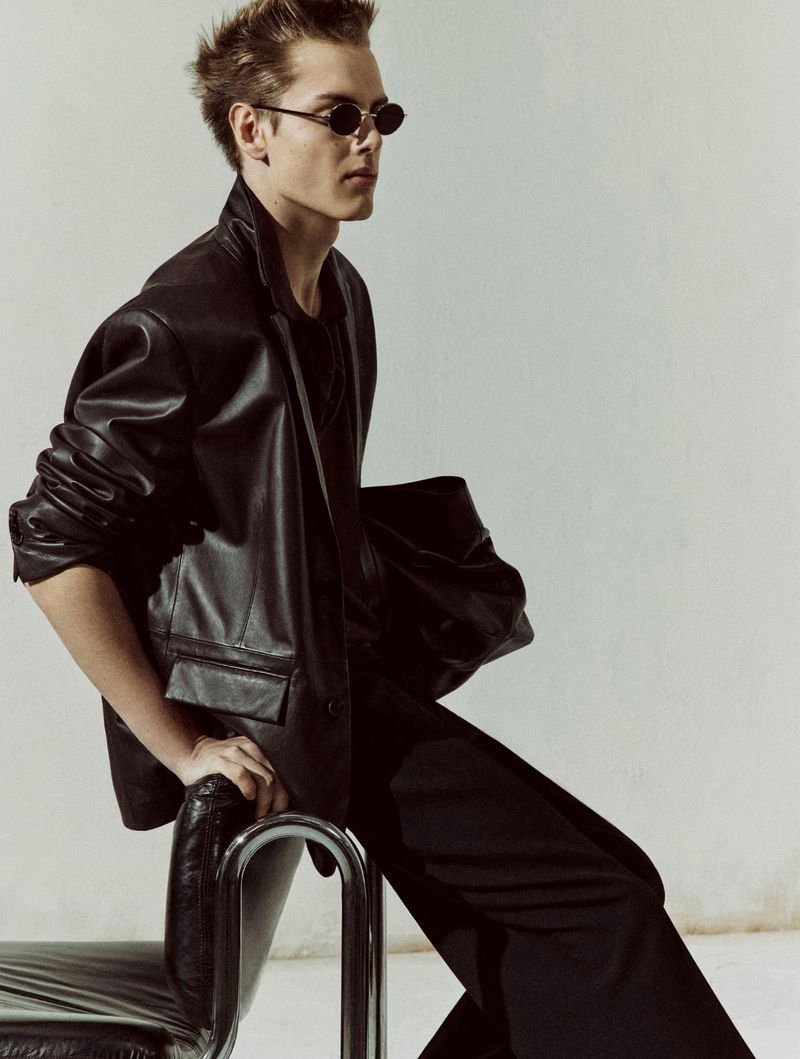 A stylish young adult in a black leather jacket and sunglasses poses on a metal chair in a fashion-forward studio shot.